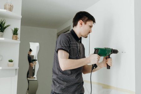 One young handsome Caucasian brunette guy stands sideways, holds a drill with both hands and drills a hole in a white wall, standing in a room and reflected behind in a figured mirror on a spring day