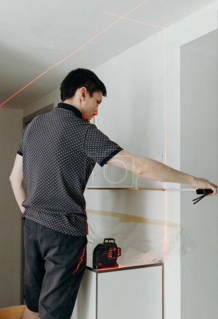 One young handsome Caucasian brunette guy stands half sideways, holds a measuring construction tape in his hands and checks the evenness of a hung wooden shelf on a white wall in a room on a spring