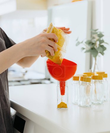 One young Caucasian unrecognizable girl pours yellow seasoning spice from a transparent bag into a glass jar while standing at a white table in the kitchen on a summer day, side view close-up with