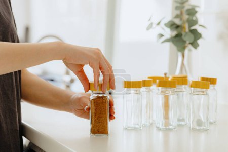One young Caucasian unrecognizable girl closes with her hands a wooden lid mix seasoning spice for fish in a glass jar, standing at a white table in the kitchen on a summer day, side view close-up
