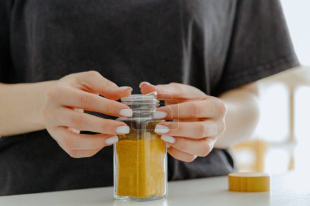 One young Caucasian unrecognizable girl closes the lid of a glass jar with curry spice while standing at a white table in the kitchen on a summer day, side view close-up with depth of field. Eco