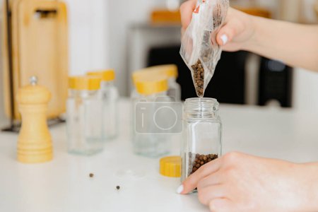 One young Caucasian unrecognizable girl pours seasoning spice black peppercorns from a plastic bag into a new glass transparent jar, standing at a white table in the kitchen on a summer day, side view