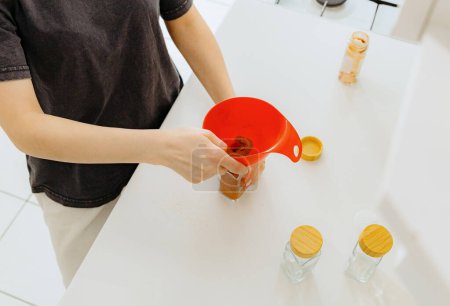 One young Caucasian unrecognizable girl pours seasoning spice ground paprika into a new glass transparent jar, standing at a white table in the kitchen on a summer day, using a red watering can, top