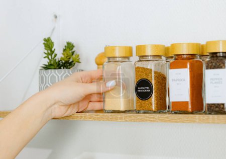One young Caucasian unrecognizable girl takes out with one hand a glass jar with the spice dry ground garlic from a wooden wall shelf in the kitchen on a summer day, side view close-up with depth of