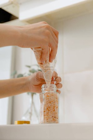 One young Caucasian unrecognizable girl pours pink salt seasoning spice from a plastic bag into a new glass transparent jar, standing at a white table in the kitchen on a summer day, bottom side view