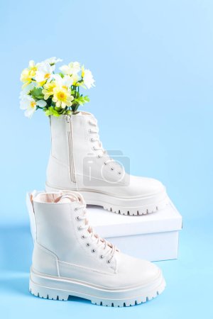 Botas demi-season martens blancas fabricadas en polipiel con suela rugosa con un ramo de flores primaverales y una caja de cartón blanco para zapatos de pie sobre fondo azul claro, vista lateral de primer plano. El