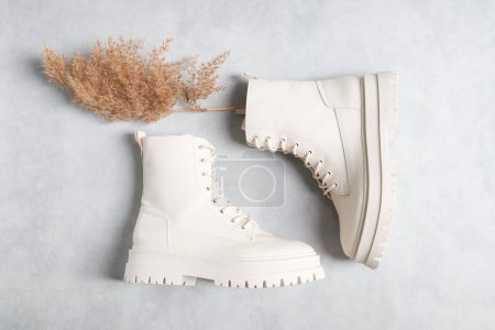 Botas blancas demi-season fabricadas en polipiel con broches, cordones, suelas ásperas y una rama de caña seca sobre un fondo de cemento gris, planas de primer plano. El concepto de moda y zapatos de mujer.