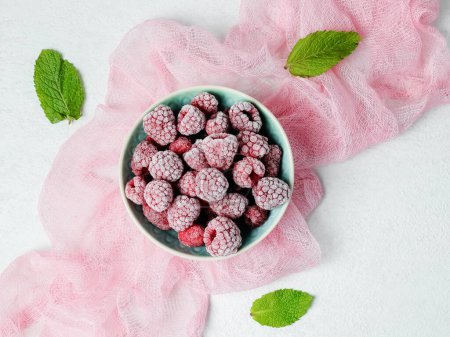 Frozen raspberries in a bowl with mint.Frozen raspberries in a bowl, three mint leaves and a pink gauze napkin lie diagonally on a light cement background, close-up top view.