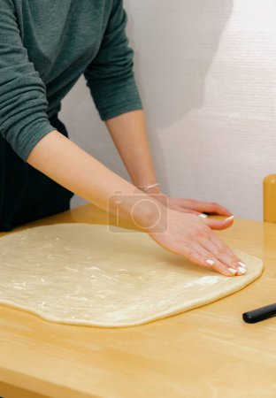 One unrecognizable young Caucasian girl spreads butter with her fingers onto the yeast dough on a wooden table, standing in the kitchen, close-up side view. Step-by-step instructions for baking
