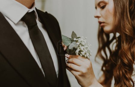 Portrait of one young beautiful stylish Caucasian brunette bride inserting a mini bouquet of boutonnieres into the grooms chest pocket in a dark suit, close-up side view. Wedding traditions concept.