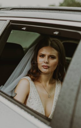 Portrait of one young beautiful stylish Caucasian brunette bride with a serious emotion on her face sits inside a car and looks at the camera through an open window on a rainy cloudy day, close-up
