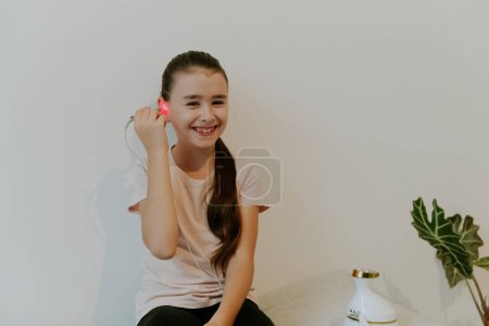 Eine schöne kaukasische Brünette mit einem glücklichen Lächeln, gesammelten Haaren und in einem rosafarbenen T-Shirt behandelt ihr Ohr mit einem Infrarotlicht-Gerät und sitzt auf ihren Knien auf einem Bett zu Hause in der Nähe einer weißen Wand