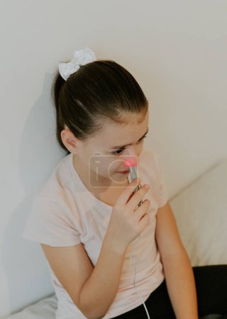 Une belle fille brune caucasienne avec une émotion grave, cheveux recueillis et dans un T-shirt rose traite le passage nasal droit avec un appareil à lumière infrarouge, assis sur le lit, jambes croisées