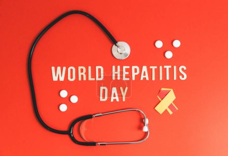 One stethoscope, white pills, a yellow-red ribbon and light wooden letters with the lined phrase World Hepatitis Day lie in the center on a red background, flat lay close-up.