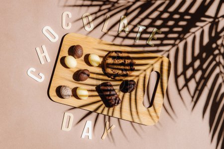 Schöne Ansicht von Schokoladenbonbons Eier und Kekse auf einem kleinen Holzbrett mit Buchstaben in der Phrase Schokolade Tag aufgereiht liegen auf einem natürlichen hellbraunen Hintergrund mit dem Schatten eines Palmenzweiges, flach lag Nahaufnahme. Konzept zum Weltschokoladentag.