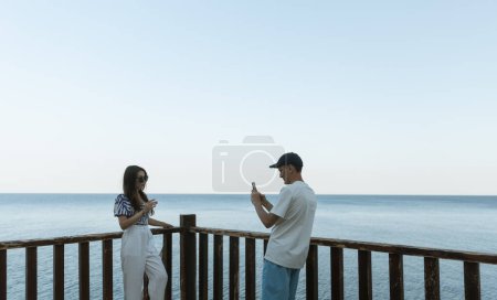 Un mec mignon dans une casquette prend une photo smartphone d'une belle fille brune en lunettes de soleil debout sur une terrasse en bois sur fond de mer lors d'une soirée d'été ensoleillée, vue latérale rapprochée.