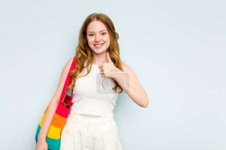 Foto de Joven mujer caucásica sosteniendo bolso LGTBI aislado sobre fondo azul sonriendo y levantando el pulgar hacia arriba - Imagen libre de derechos