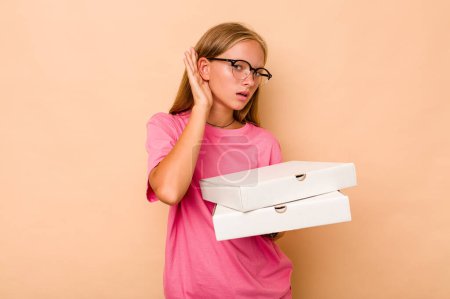 Téléchargez les photos : Petite fille caucasienne tenant une pizza isolée sur fond beige essayant d'écouter un potins. - en image libre de droit