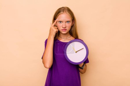 Foto de Niña caucásica sosteniendo un reloj aislado sobre fondo beige mostrando un gesto de decepción con el dedo índice. - Imagen libre de derechos