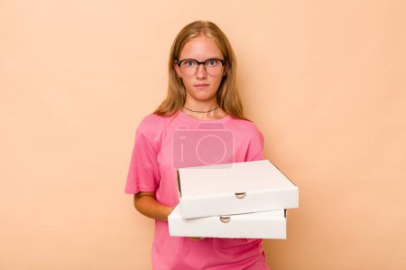 Foto de Pequeña chica caucásica sosteniendo pizza aislada sobre fondo beige encoge hombros y ojos abiertos confundidos. - Imagen libre de derechos