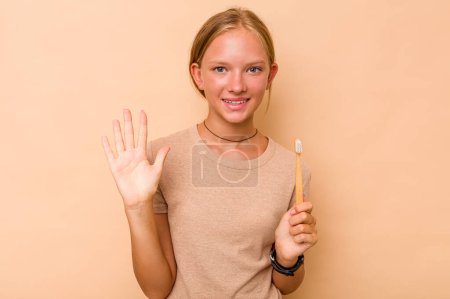 Foto de Caucásico adolescente chica cepillarse los dientes aislado en beige fondo sonriendo alegre mostrando número cinco con los dedos. - Imagen libre de derechos