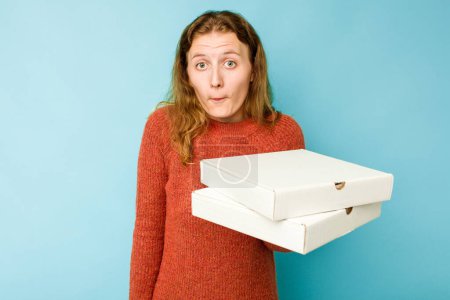 Photo for Young caucasian woman holding pizzas boxes isolated on blue background shrugs shoulders and open eyes confused. - Royalty Free Image