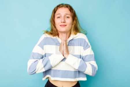 Foto de Joven mujer caucásica aislada sobre fondo azul cogida de la mano en orar cerca de la boca, se siente segura. - Imagen libre de derechos