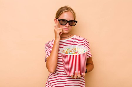Photo for Little caucasian girl eating popcorn isolated on beige background pointing temple with finger, thinking, focused on a task. - Royalty Free Image