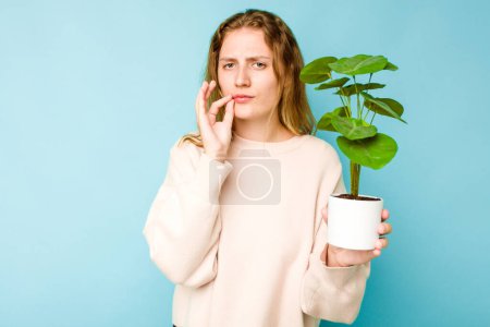 Foto de Mujer caucásica joven sosteniendo una planta aislada sobre fondo azul con los dedos en los labios manteniendo un secreto. - Imagen libre de derechos
