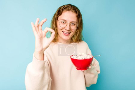 Photo for Young caucasian woman holding a bowl of cereals isolated on blue background cheerful and confident showing ok gesture. - Royalty Free Image