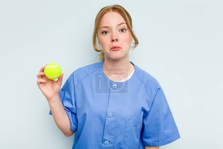 Téléchargez les photos : Jeune kinésithérapeute caucasien tenant une balle de tennis isolée sur fond bleu haussant les épaules et ouvrant les yeux confus. - en image libre de droit