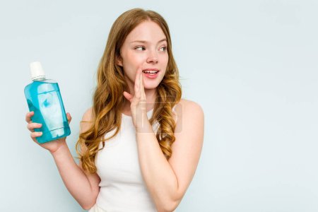 Photo for Young caucasian woman holding a mouthwash isolated on blue background is saying a secret hot braking news and looking aside - Royalty Free Image