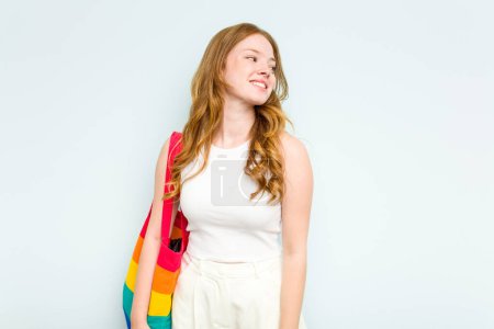 Foto de Joven mujer caucásica sosteniendo LGTBI bolso aislado sobre fondo azul mira a un lado sonriente, alegre y agradable. - Imagen libre de derechos