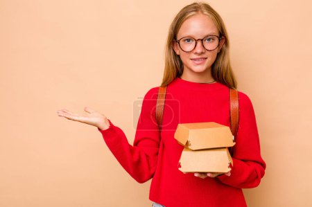 Photo for Little caucasian student girl holding burgers isolated on beige background showing a copy space on a palm and holding another hand on waist. - Royalty Free Image