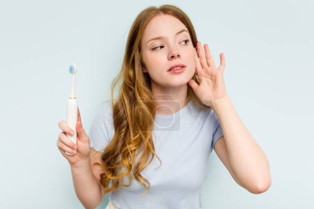 Téléchargez les photos : Jeune femme caucasienne tenant une brosse à dents électrique isolée sur fond bleu essayant d'écouter un potins. - en image libre de droit