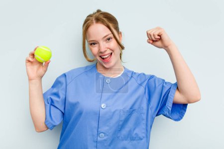Photo for Young caucasian physiotherapist holding a tennis ball isolated on blue background raising fist after a victory, winner concept. - Royalty Free Image