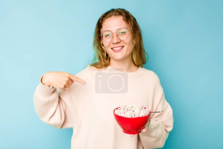 Foto de Mujer caucásica joven sosteniendo un tazón de cereales aislado sobre fondo azul persona señalando a mano a un espacio de copia de la camisa, orgulloso y confiado - Imagen libre de derechos