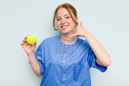 Téléchargez les photos : Jeune kinésithérapeute caucasien tenant une balle de tennis isolée sur fond bleu montrant un geste d'appel téléphonique mobile avec les doigts. - en image libre de droit