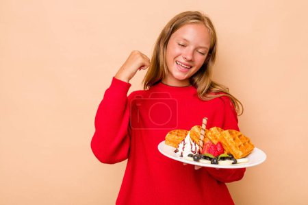 Photo for Little caucasian girl holding a waffles isolated on beige background raising fist after a victory, winner concept. - Royalty Free Image