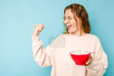 Téléchargez les photos : Jeune femme caucasienne tenant un bol de céréales isolé sur fond bleu levant le poing après une victoire, concept gagnant. - en image libre de droit