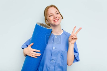Photo for Young caucasian physiotherapist holding a mat isolated on blue background joyful and carefree showing a peace symbol with fingers. - Royalty Free Image