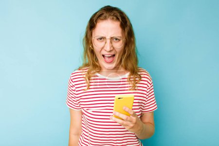 Photo for Young caucasian woman holding mobile phone isolated on blue background screaming very angry and aggressive. - Royalty Free Image