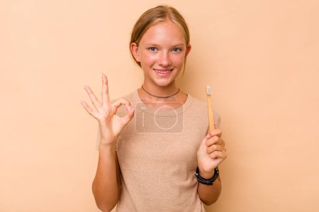 Téléchargez les photos : Caucasien adolescent fille brossage dents isolé sur beige fond gai et confiant montrant ok geste. - en image libre de droit