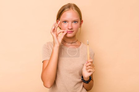 Téléchargez les photos : Caucasien adolescent fille brossage dents isolé sur fond beige avec les doigts sur les lèvres garder un secret. - en image libre de droit