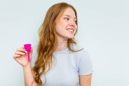 Téléchargez les photos : Jeune femme caucasienne tenant la coupe menstruelle isolée sur fond bleu regarde de côté souriant, gai et agréable. - en image libre de droit