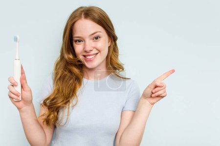 Téléchargez les photos : Jeune femme caucasienne tenant une brosse à dents électrique isolée sur fond bleu souriant et pointant du doigt, montrant quelque chose à vide. - en image libre de droit