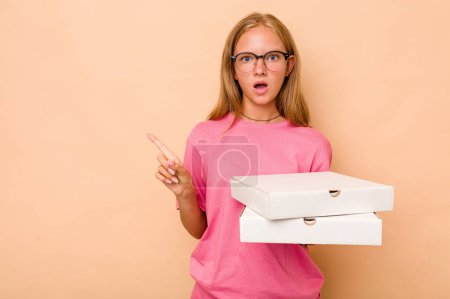 Téléchargez les photos : Petite fille caucasienne tenant la pizza isolée sur fond beige pointant vers le côté - en image libre de droit