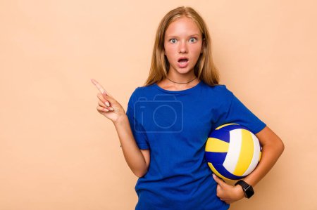 Téléchargez les photos : Petite fille caucasienne jouant au volley isolé sur fond beige pointant vers le côté - en image libre de droit