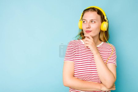 Téléchargez les photos : Jeune femme caucasienne portant des écouteurs isolés sur fond bleu regardant latéralement avec une expression douteuse et sceptique. - en image libre de droit