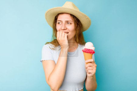 Foto de Mujer caucásica joven sosteniendo un helado aisló un fondo azul mordiendo las uñas, nervioso y muy ansioso. - Imagen libre de derechos